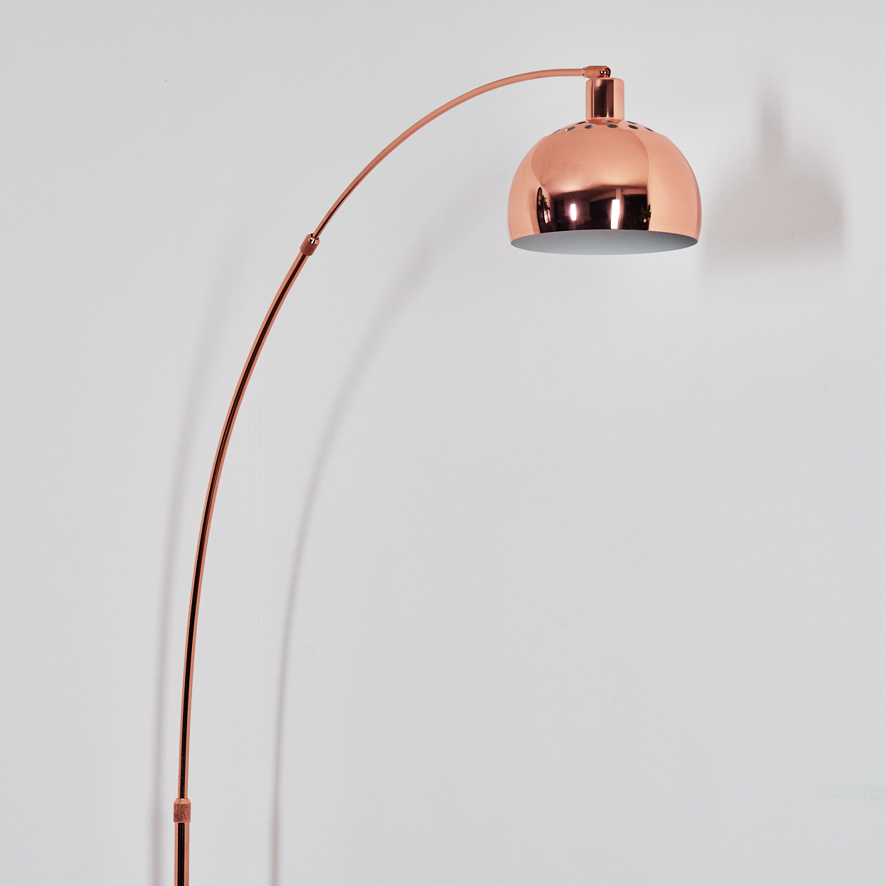 Eine kupferfarbene Bogenlampe steht vor einer weißen Wand. Die Lampe hat einen großen, runden Schirm und einen gebogenen Arm.