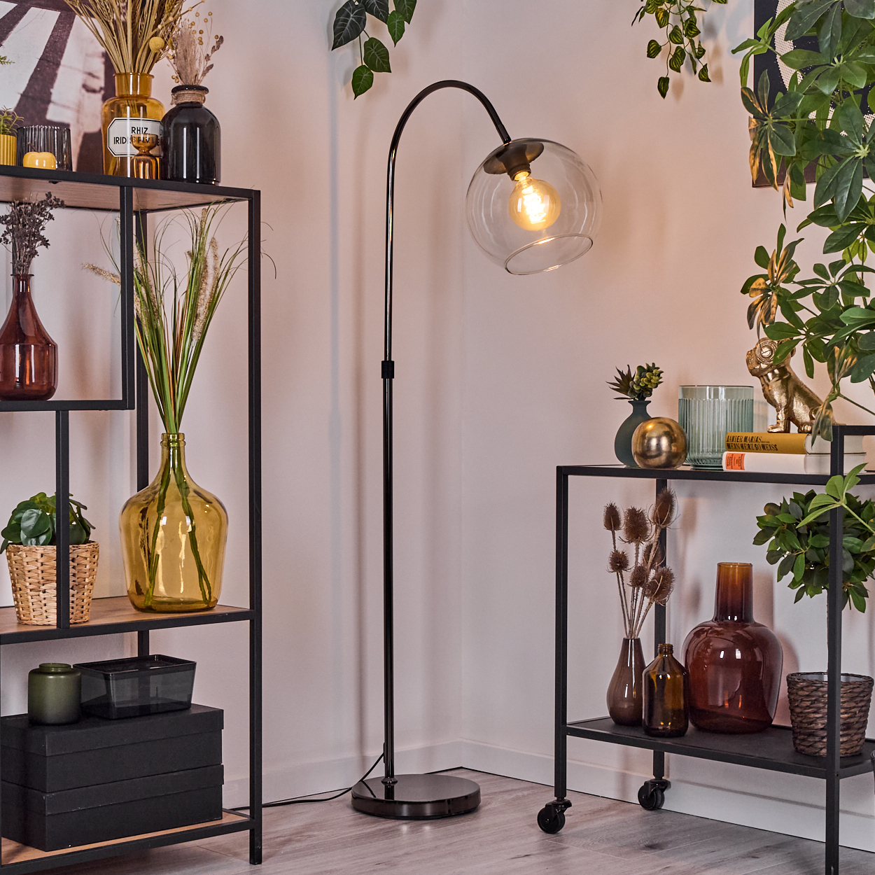 Innenaufnahme mit Stehlampe. Regale mit Vasen, Pflanzen und Dekor. Grün hängt von der Decke. Eine goldene Hundestatue ist sichtbar.