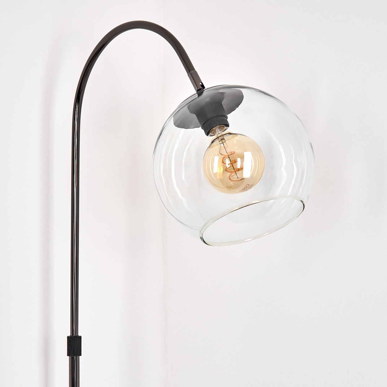 Eine moderne Stehlampe mit dunklem Metallrahmen und Glaskugelschirm. Die Glühbirne im Inneren leuchtet vor einer weißen Wand.