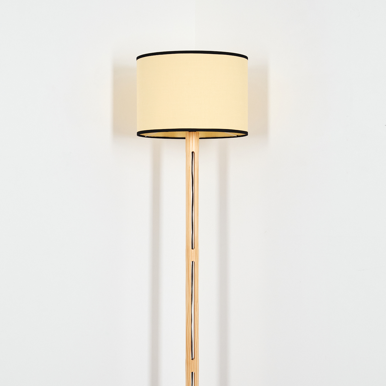 Eine Stehlampe mit beigem Lampenschirm und Holzfuß. Der Lampenschirm hat eine schwarze Einfassung. Der Hintergrund ist eine weiße Wand.