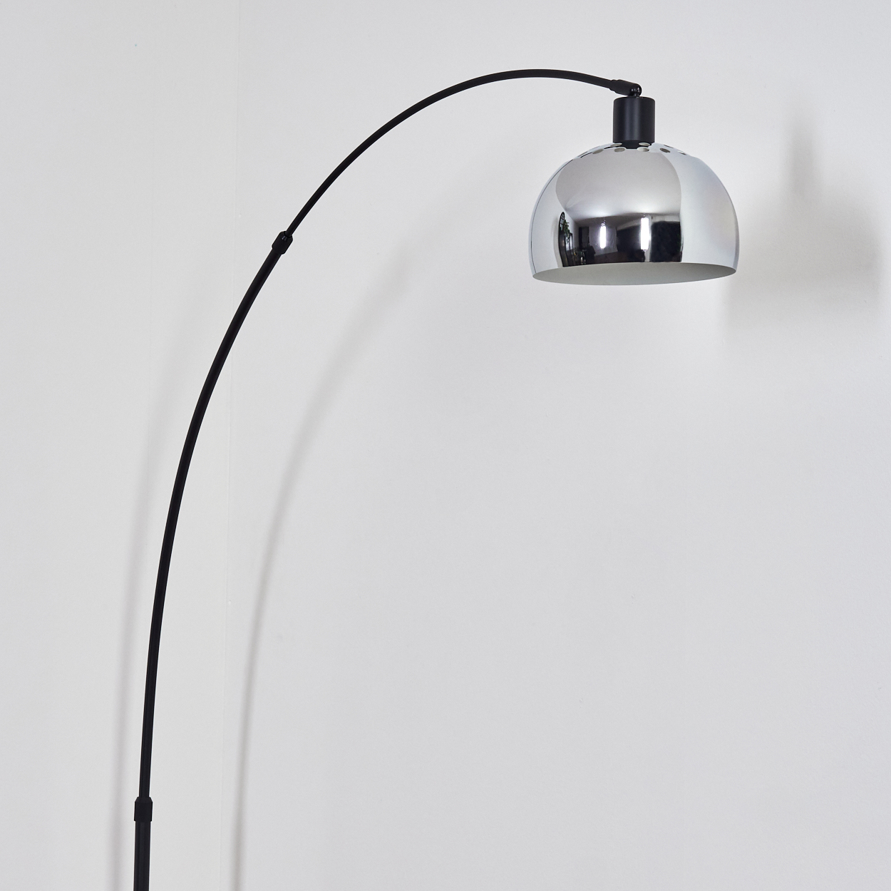 Eine moderne Bogen-Stehlampe mit verchromtem Lampenschirm und schwarzem, gebogenem Arm. Der Hintergrund ist eine schlichte, weiße Wand.