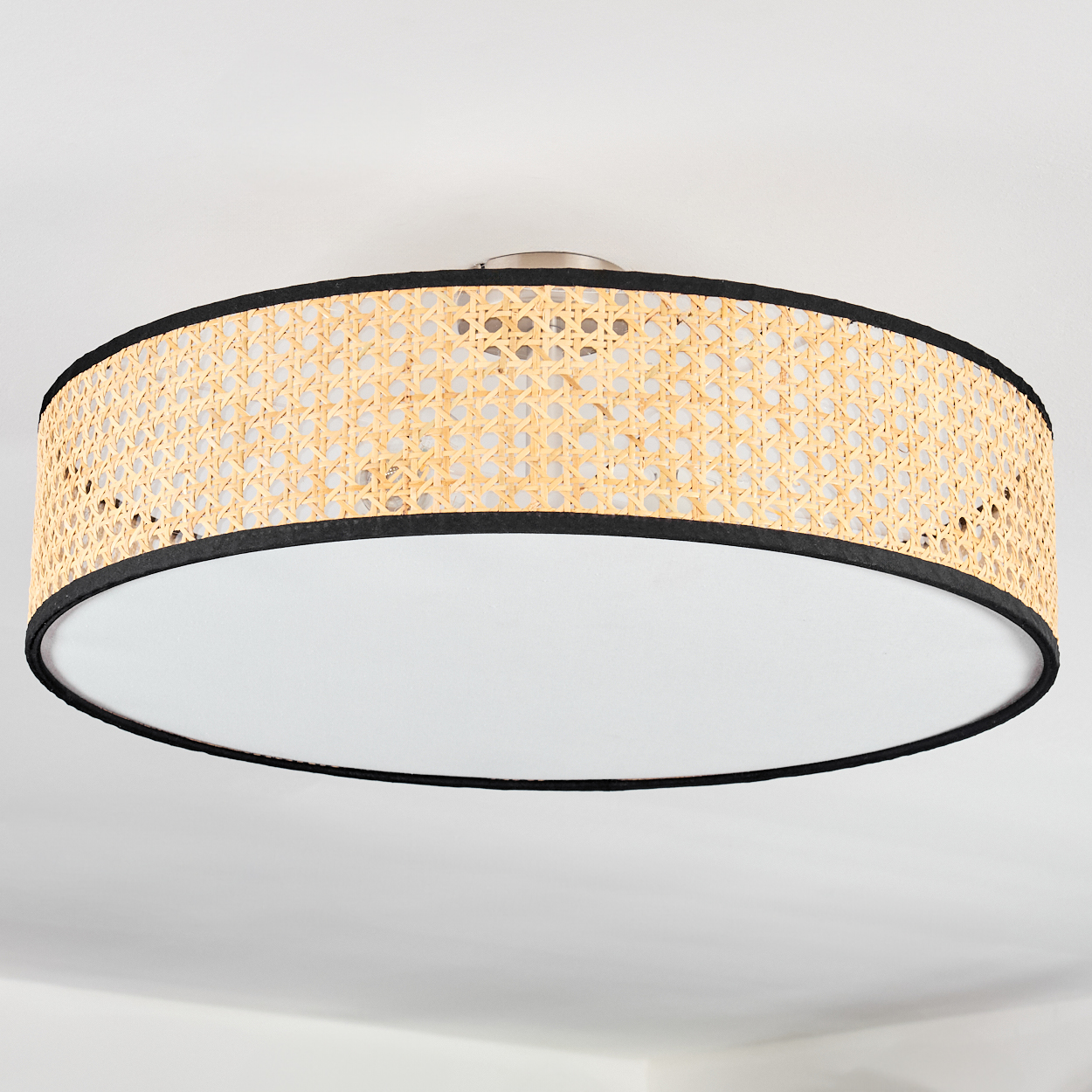 Eine runde Deckenlampe. Sie hat einen hellen, gewebten Holzschirm. Der Schirm ist schwarz eingefasst. Der Boden ist weiß.