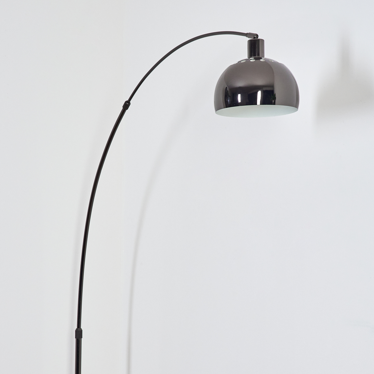 Eine moderne, schwarze Bogen-Stehlampe mit einem metallisch-grauen Lampenschirm steht vor einer weißen Wand. Ein Schatten wird auf die Wand geworfen.