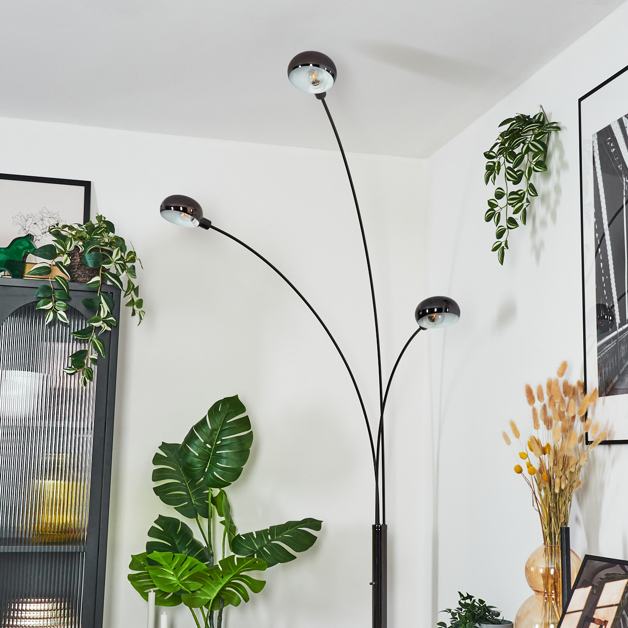 Eine moderne Stehlampe mit drei schwarzen, gebogenen Armen. Pflanzen, ein Bild und Dekorationsartikel sind im Hintergrund. Weiße Wände.