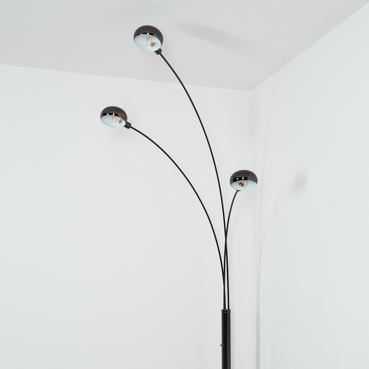 Eine moderne schwarze Stehlampe mit drei gebogenen Armen und kugelförmigen Lampenschirmen, vor einer weißen Wand in einer Ecke.