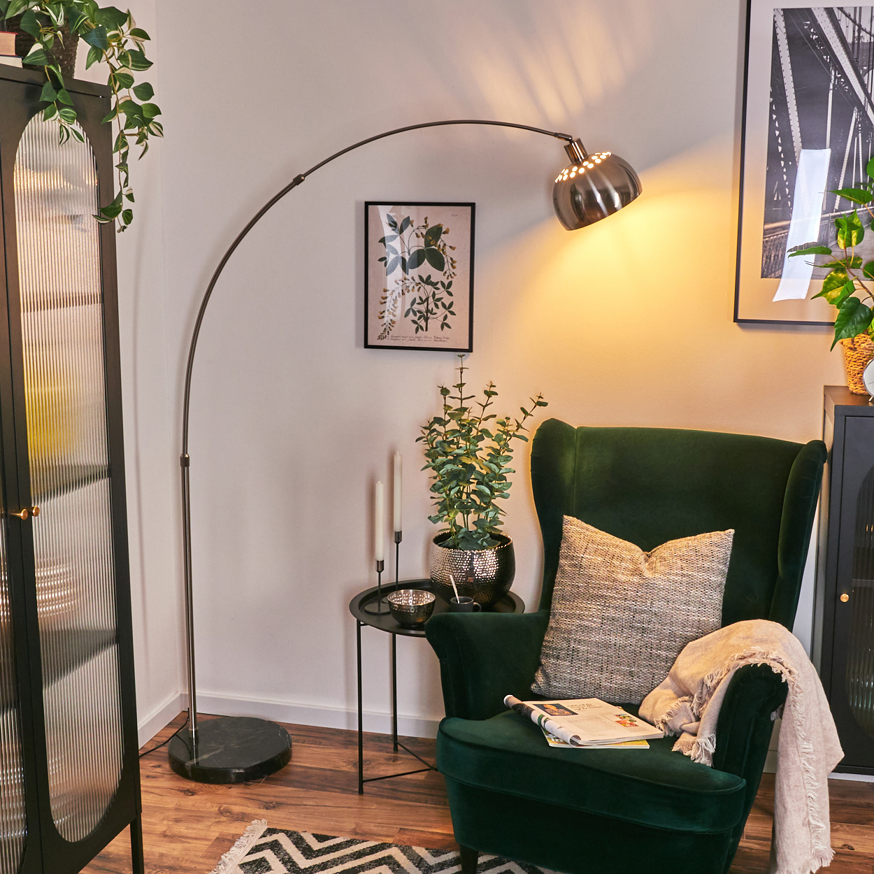 Ein dunkelgrüner Sessel mit Kissen und Decke. Eine Lampe beleuchtet die Szene mit Pflanzen und Dekoration.