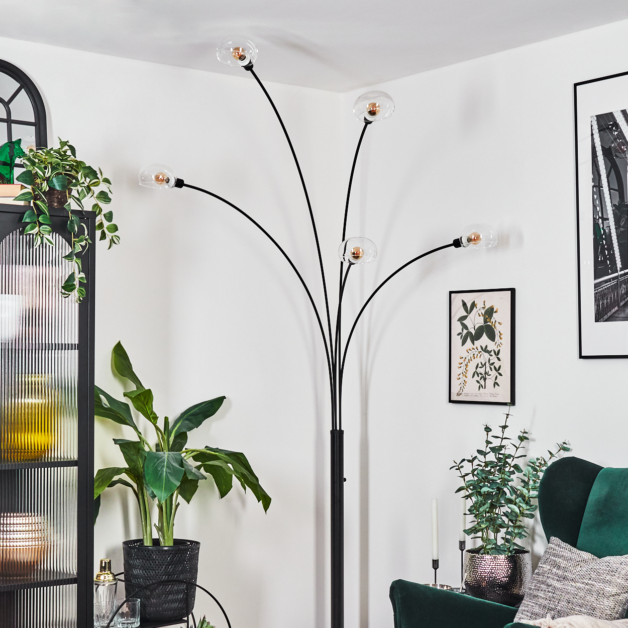 Ein modernes Wohnzimmer mit schwarzer Stehlampe, Pflanzen, grünem Sessel und gerahmter Kunst. Weiße Wände und schwarzes Regal.