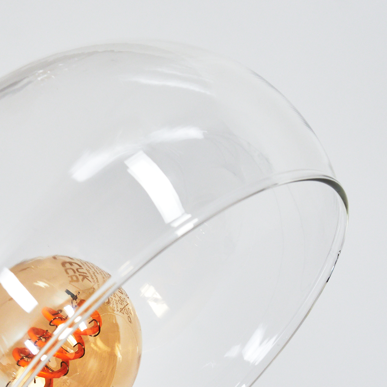 Nahaufnahme eines Glasschirms, der eine Glühbirne umschließt. Der Glühfaden der Birne leuchtet orange, der Schirm ist klar, vor weißem Hintergrund.