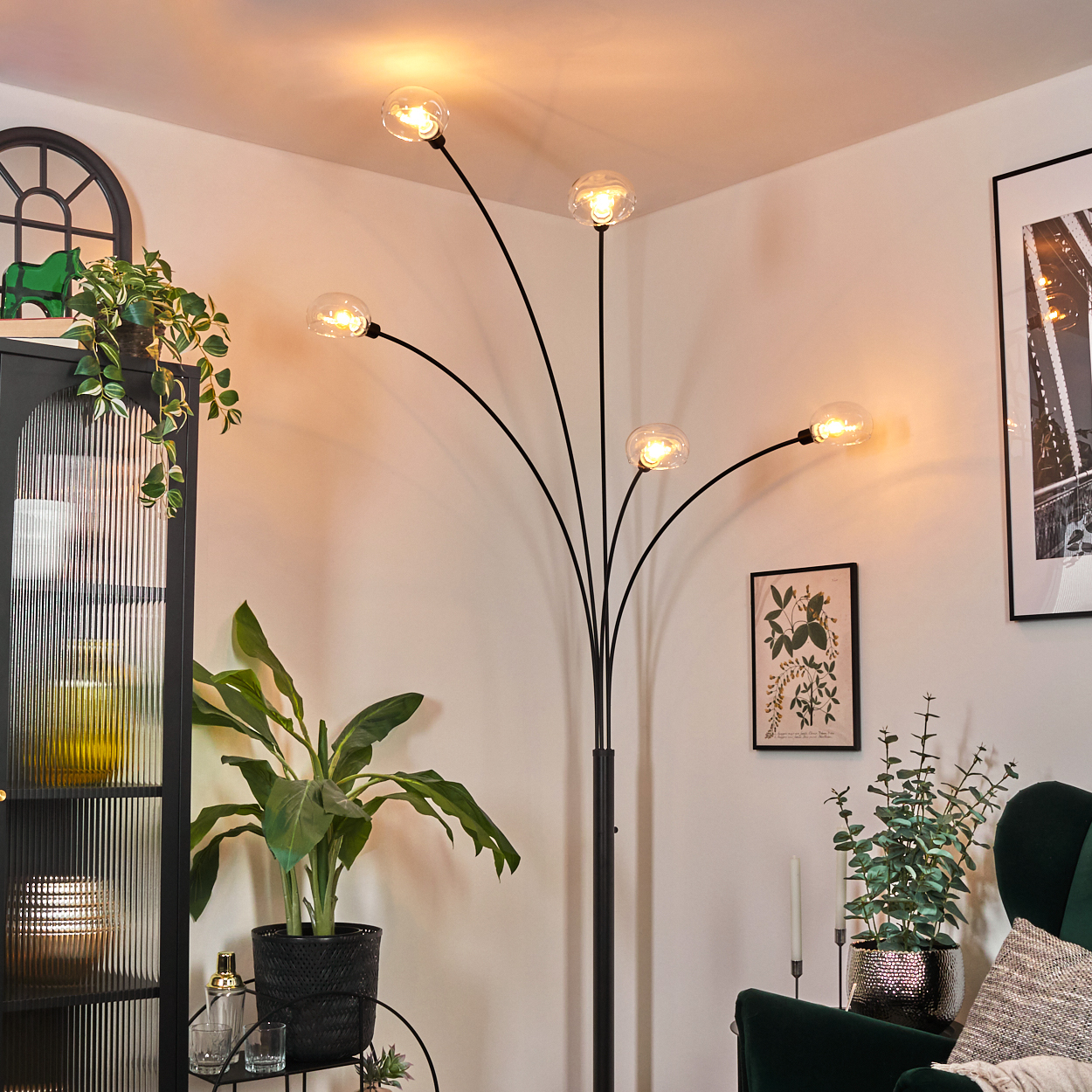 Ein modernes Wohnzimmer mit einer schwarzen Stehlampe mit fünf Lichtern. Pflanzen und Bilder schmücken den Raum. Ein grüner Sessel ist sichtbar.