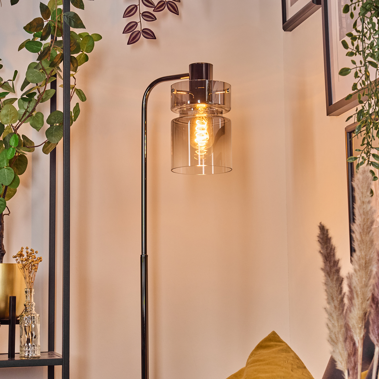 Eine moderne Stehlampe mit Glasschirm und sichtbarer Glühbirne. Grüne Pflanzen und ein dekoratives Regal im Hintergrund.