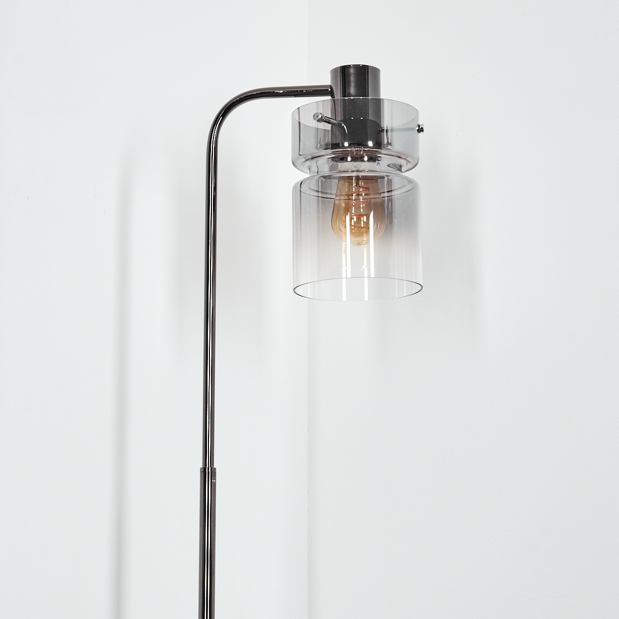 Eine moderne Stehlampe mit gebogenem Arm, der einen zylindrischen Glasschirm hält. Die Glühbirne im Inneren ist sichtbar, der Hintergrund ist weiß.