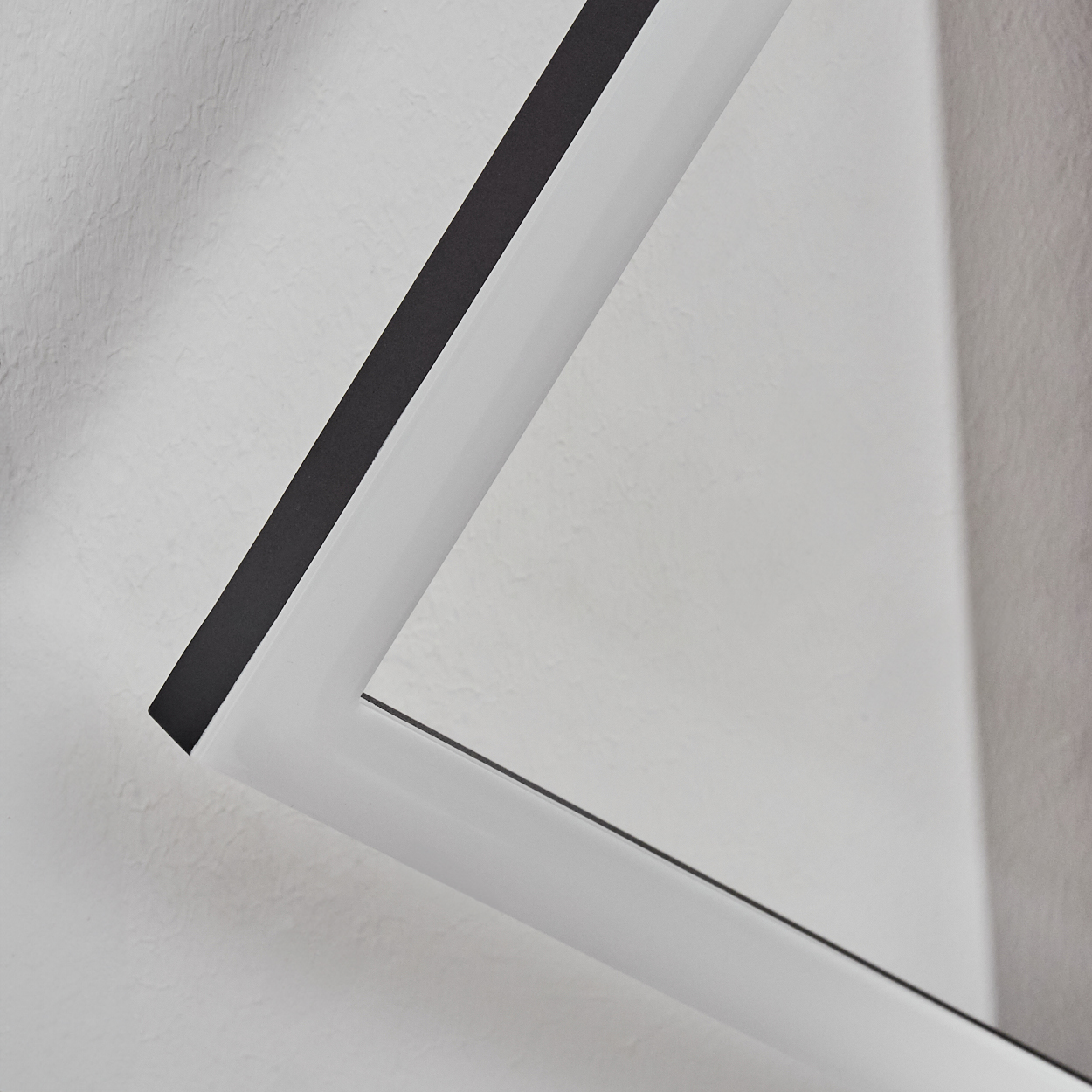 Nahaufnahme eines weißen Rahmens mit schwarzem Innenrand vor einer strukturierten weißen Wand. Der Rahmen bildet eine geometrische Form.