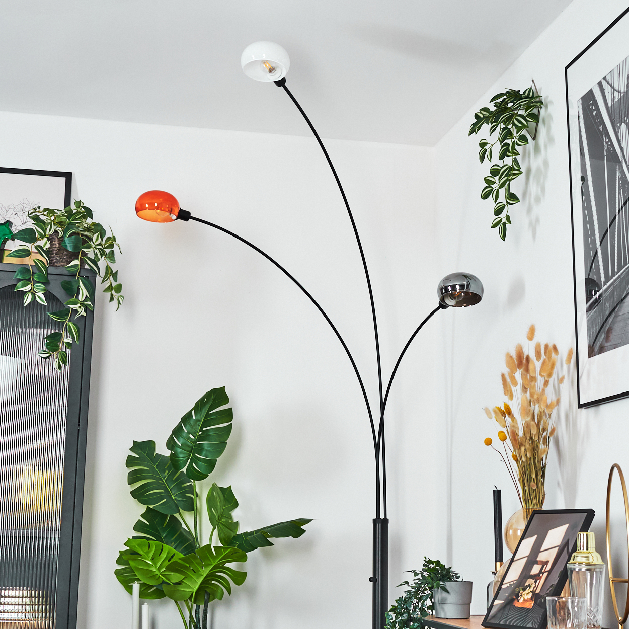 Eine moderne Stehlampe mit drei Köpfen, jede mit einem anderen Farbton, steht in einem Raum mit Pflanzen und Dekorationen.