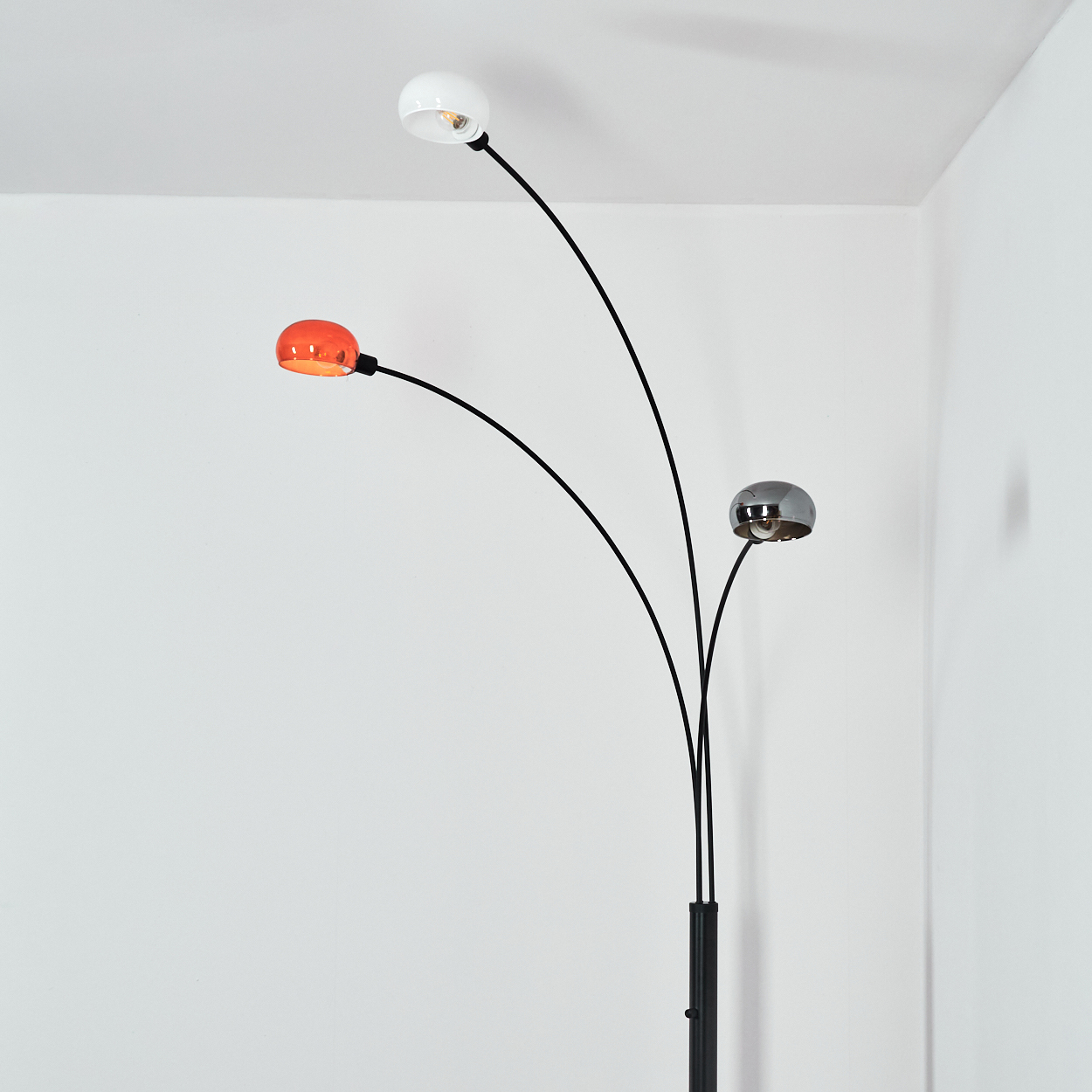 Eine moderne Stehlampe mit drei gebogenen Armen. Jeder Arm hält ein andersfarbiges Licht: weiß, orange und silber. Vor weißen Wänden.