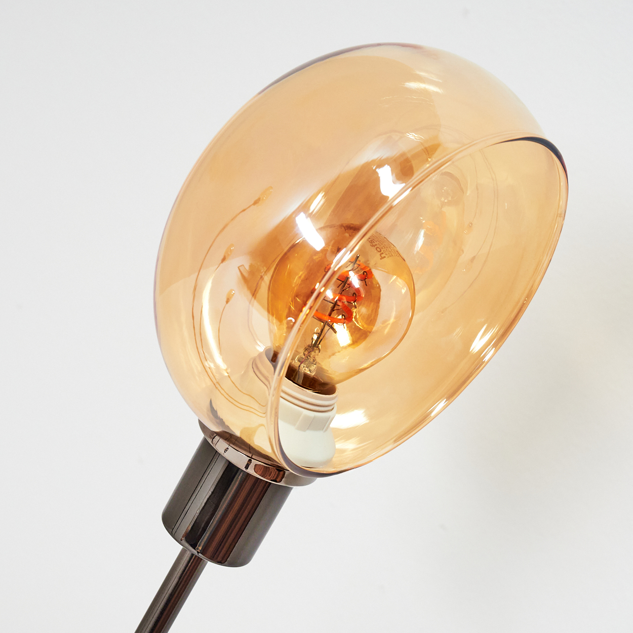 Nahaufnahme einer Lampe. Sie hat einen gebogenen, bernsteinfarbenen Glasschirm. Die Glühbirne ist sichtbar. Der Sockel ist dunkel metallisch, vor weißem Hintergrund.