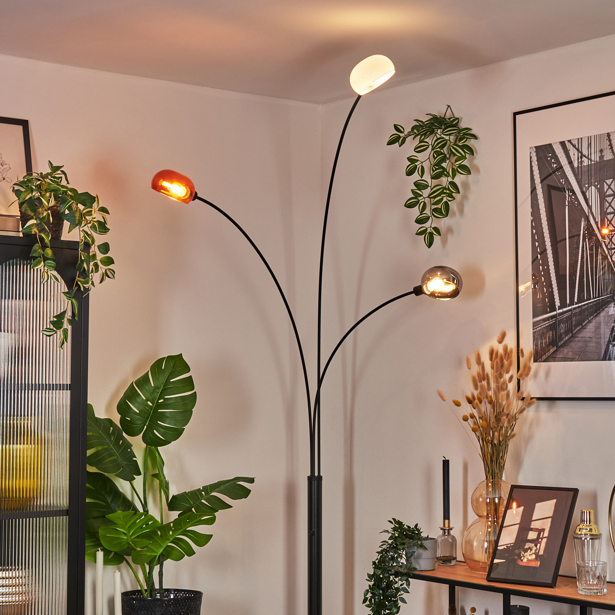 Eine moderne Stehlampe mit drei Lichtern. Pflanzen und Bilder dekorieren den Raum. Warmes Licht beleuchtet die Szene.