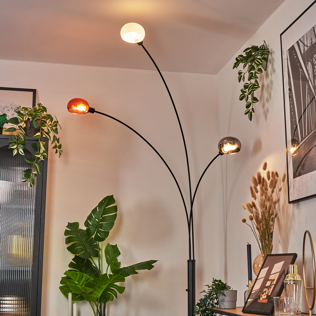 Eine moderne Stehlampe mit drei Lichtern. Vor der Lampe befindet sich eine große grüne Pflanze. Pflanzen und Kunstwerke sind im Hintergrund.