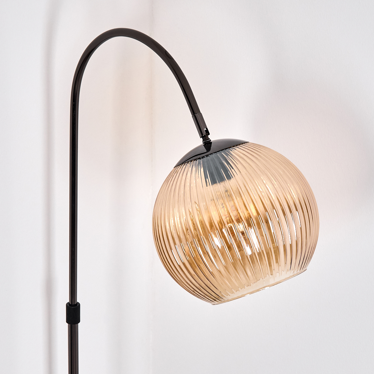 Eine moderne Stehlampe mit gebogenem schwarzem Stiel und einem kugelförmigen, gerippten bernsteinfarbenen Glasschirm vor weißer Wand.