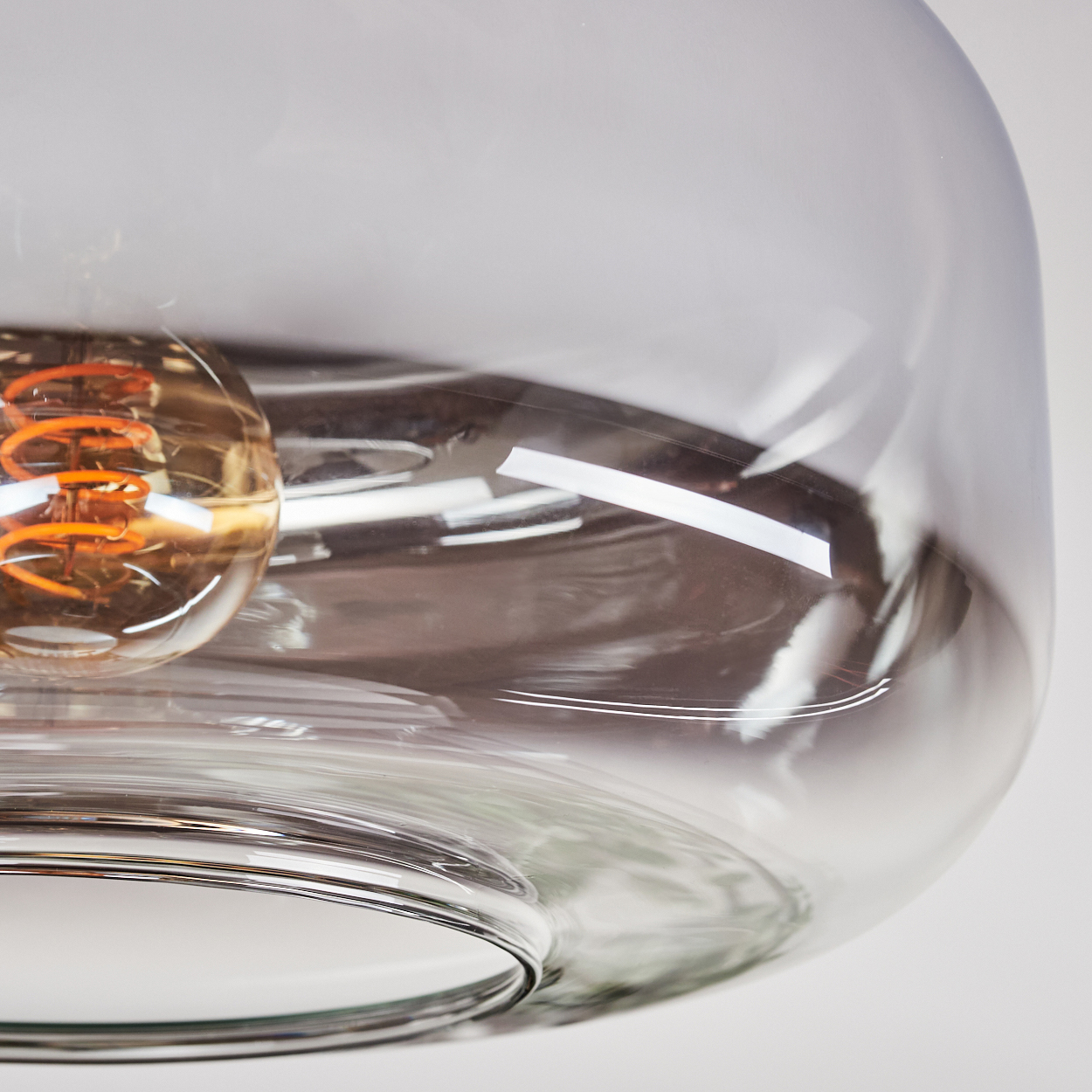 Nahaufnahme einer Glasleuchte. Ein klarer Glasschirm umgibt eine leuchtende, orangefarbene Glühbirne, der Hintergrund ist weiß.