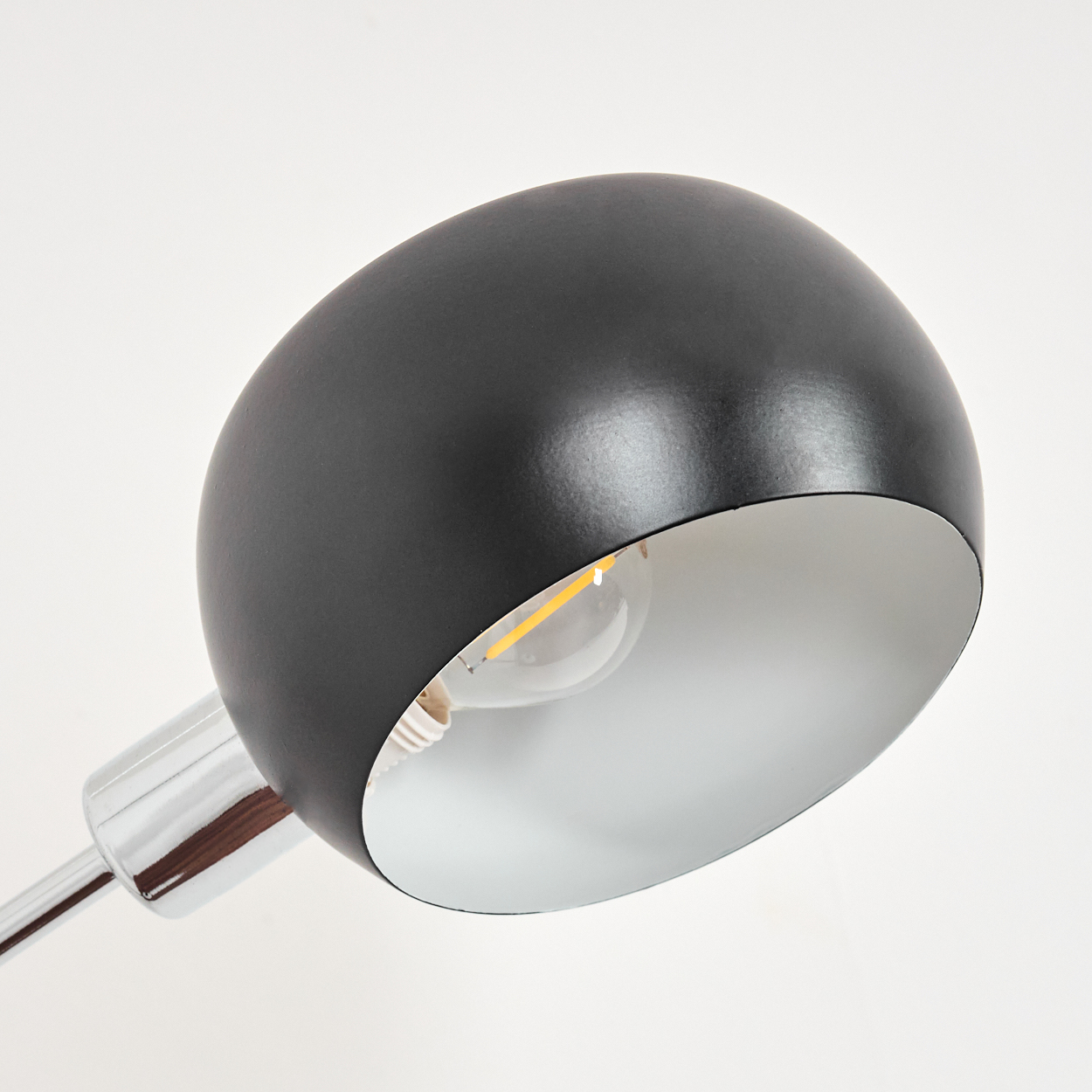 Nahaufnahme eines schwarzen Lampenkopfs mit silbernem Innenraum und sichtbarer Glühbirne. Die Lampe befindet sich auf einem Chromarm, vor weißem Hintergrund.