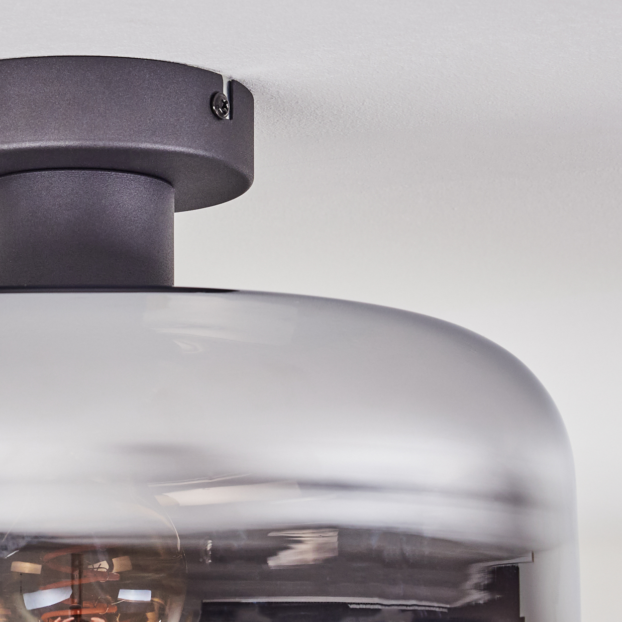 Nahaufnahme einer Deckenlampe. Sie hat einen Rauchglasschirm und einen dunkelgrauen Metallsockel. Der Hintergrund ist eine weiße Decke.