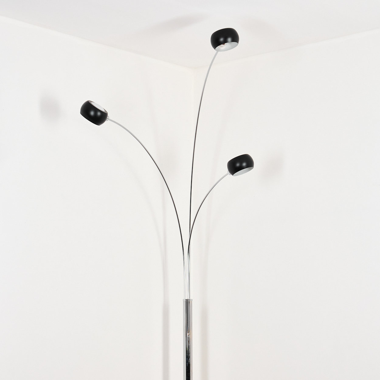Eine moderne Stehlampe mit drei schwarzen Lampenschirmen an gebogenen Stielen. Der Sockel ist verchromt, vor einer weißen Wand.