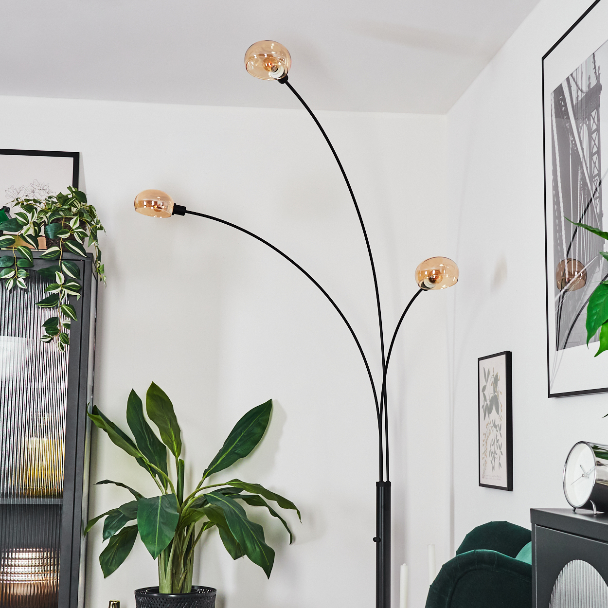 Eine moderne Stehlampe mit drei bernsteinfarbenen Glühbirnen. Grüne Pflanzen in Töpfen und ein schwarzer Schrank im Hintergrund.