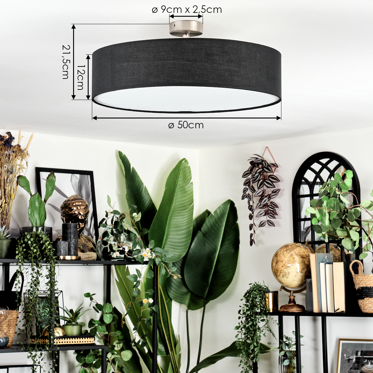 Eine schwarze zylindrische Deckenlampe ist zentriert, mit Beschriftung der Abmessungen. Darunter zeigt ein Regal Pflanzen, einen Globus und gerahmte Kunst vor einer weißen Wand.