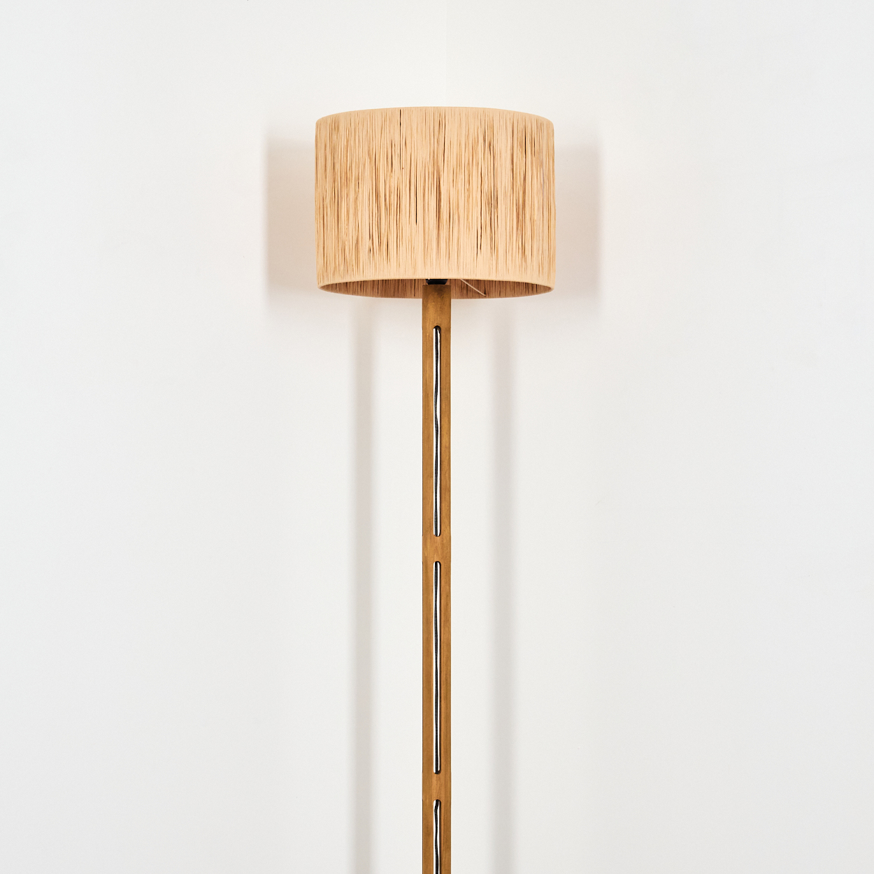 Ein beigefarbener Lampenschirm sitzt auf einem hölzernen Lampenständer. Die Lampe steht in einer Ecke an einer weißen Wand.