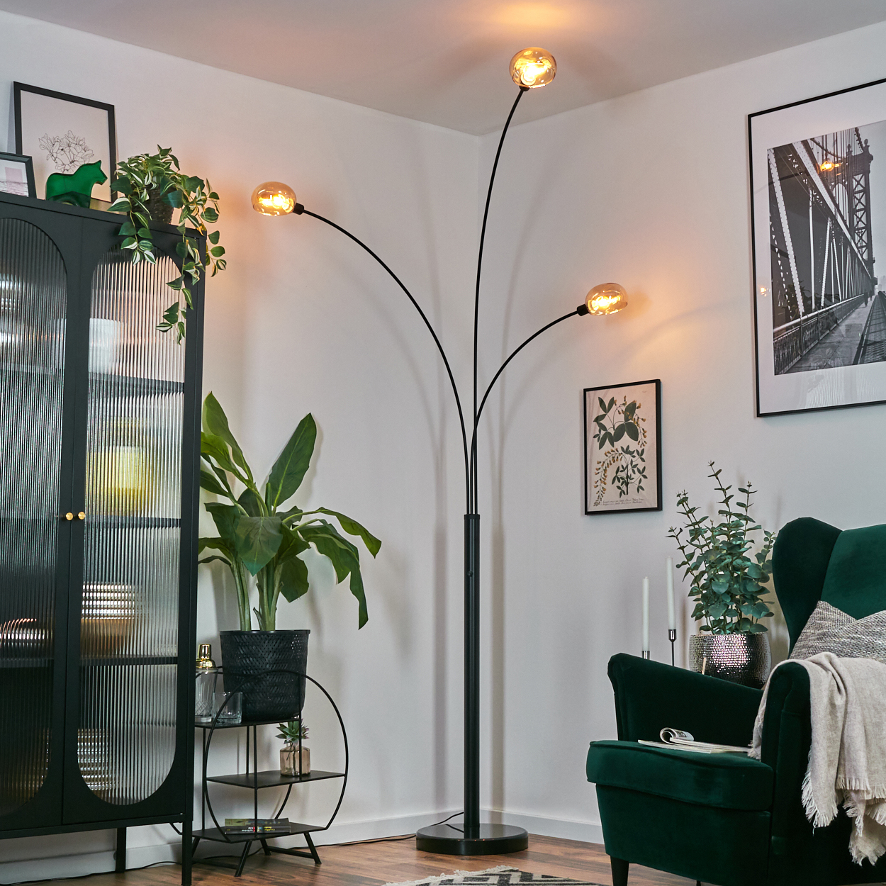 Ein modernes Wohnzimmer mit schwarzer Lampe, grünem Sessel und Pflanzen. Ein Brückenbild hängt an der Wand.
