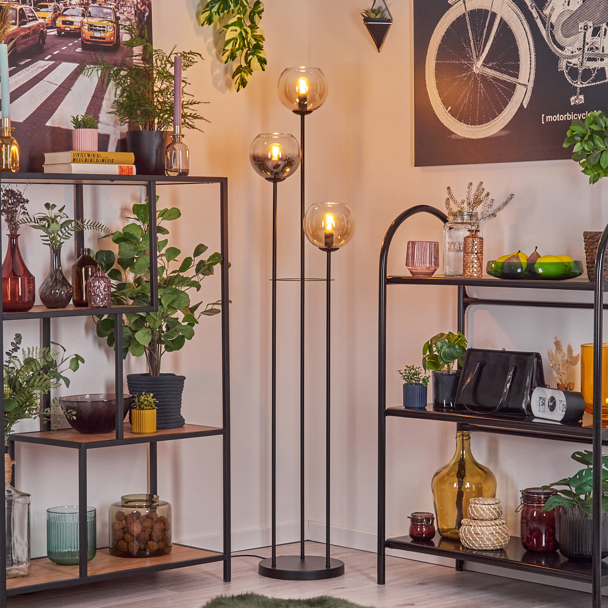 Wohnzimmer mit Regalen, Lampen und Pflanzen. Ein Fahrradkunstwerk und ein Straßenszenenbild sind ebenfalls zu sehen.