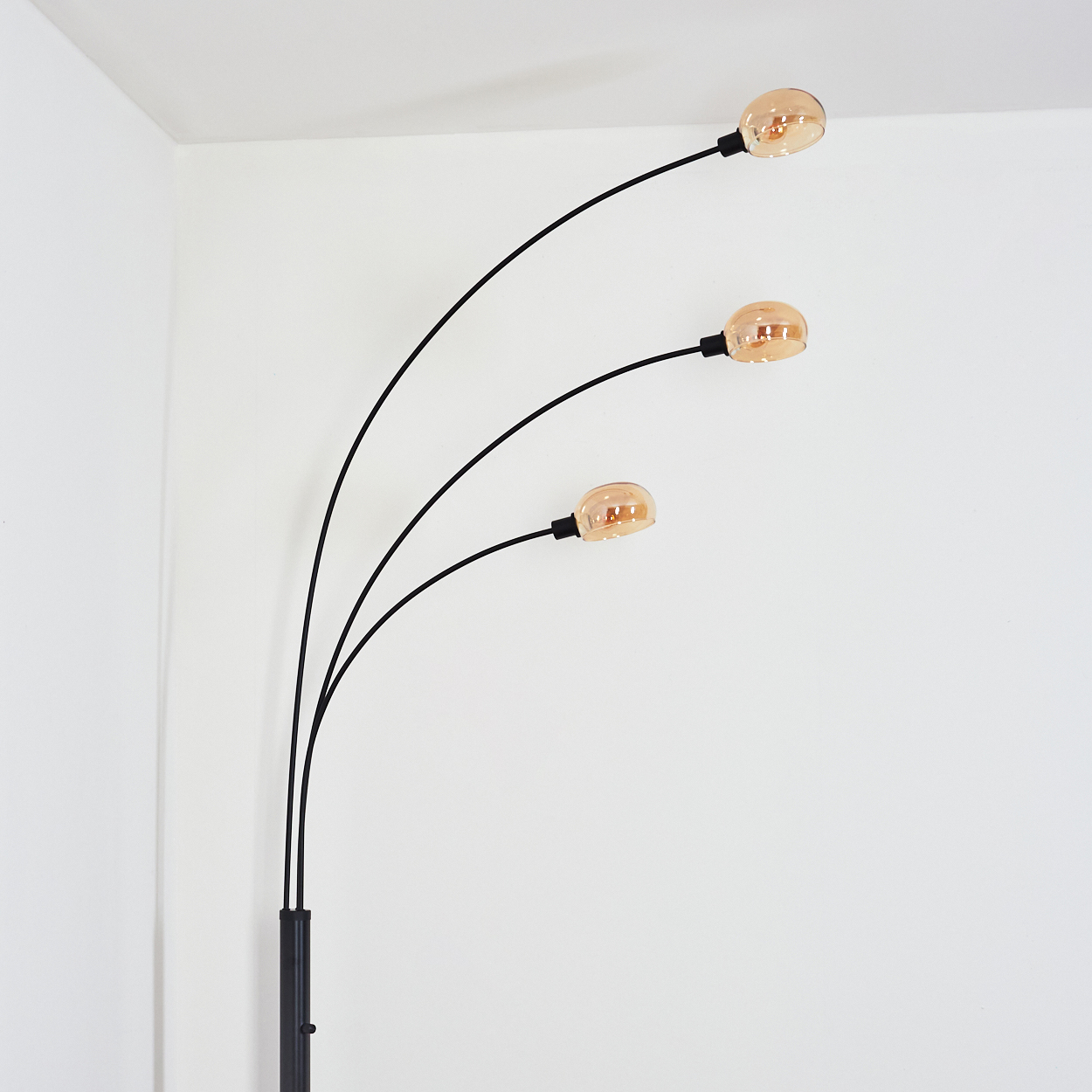 Eine schwarze Bogen-Stehlampe mit drei Glühbirnen steht vor einer weißen Wand. Die Glühbirnen sind orange und die Lampe steht in einer Ecke.