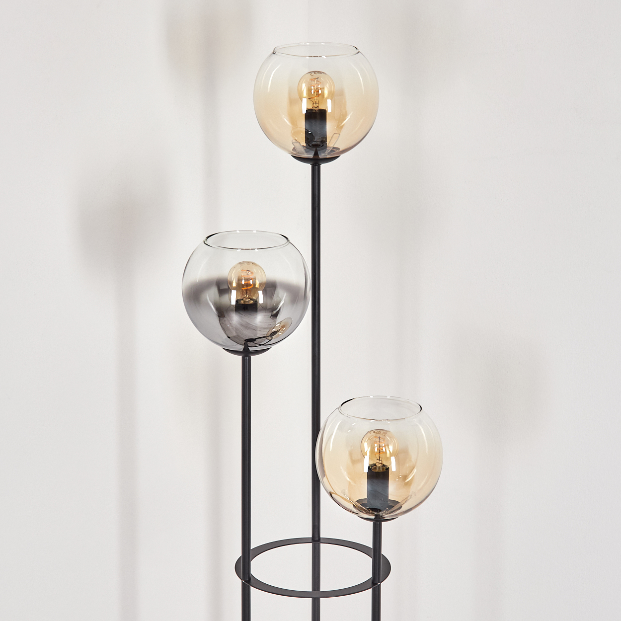 Eine moderne Stehlampe mit drei Glaskugelschirmen, die jeweils eine Glühbirne enthalten. Die Lampe hat ein schwarzes Metallgestell vor einer weißen Wand.