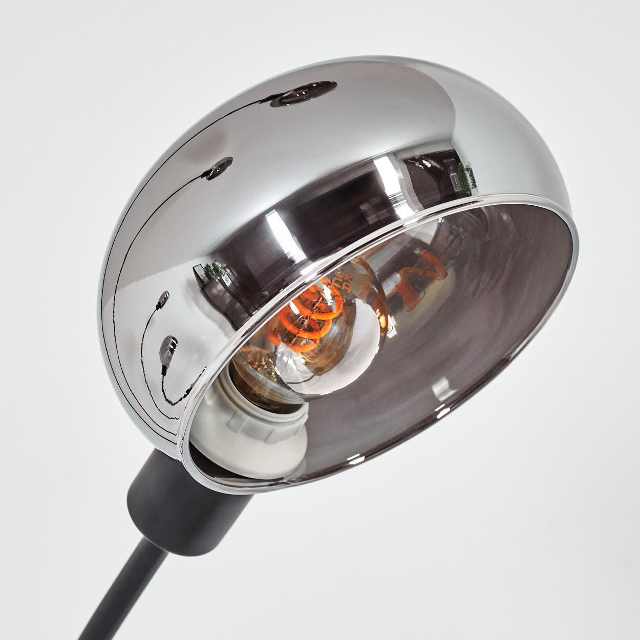 Eine Chromlampe mit sichtbarer Glühbirne. Die Lampe hat einen schwarzen Stiel und befindet sich vor weißem Hintergrund.