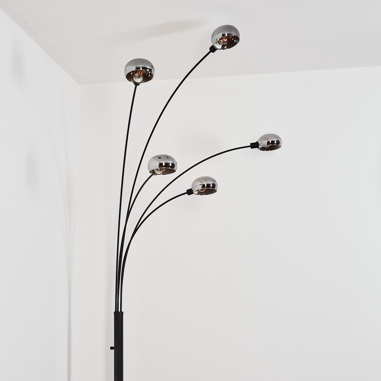 Eine moderne Stehlampe mit fünf verchromten Kugelleuchten an schwarzen Stielen vor einer weißen Wand und Decke. Die Lampe steht in der Ecke.