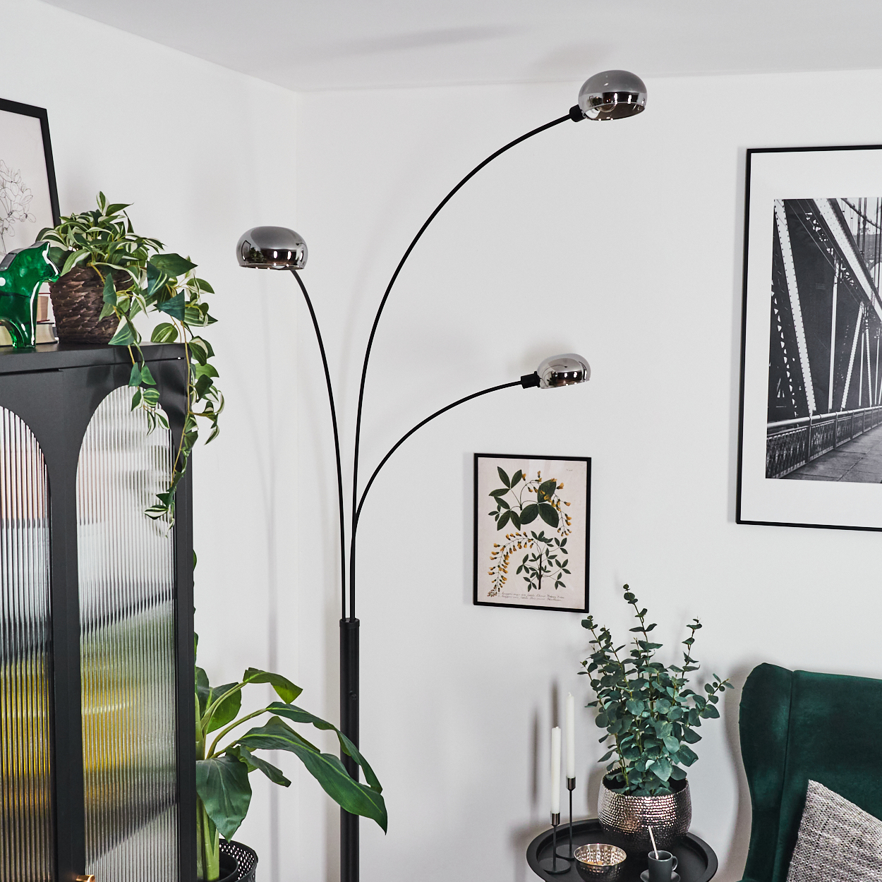 Wohnzimmer mit schwarzer Lampe, Pflanzen, Kunstwerken und einem grünen Sessel. Die Wände sind weiß.