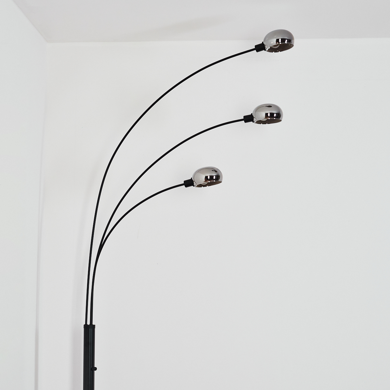 Eine moderne schwarze Stehlampe mit drei verchromten Lampen. Sie steht in einer Ecke vor einer weißen Wand.