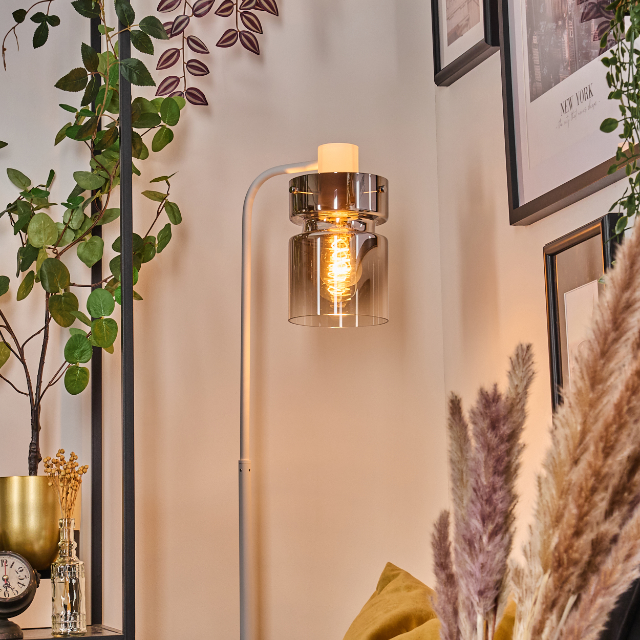 Eine beleuchtete Lampe mit Glasschirm, Pflanzen und Wandkunst schmücken eine Zimmerecke. Eine Uhr und Blumen stehen auf einem Regal.