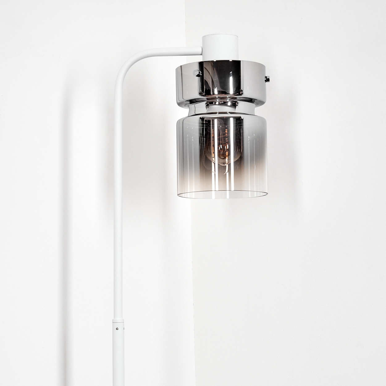 Eine moderne Lampe mit Chrom- und Glasschirm, hängend an einem weißen Arm, vor einer weißen Wand.