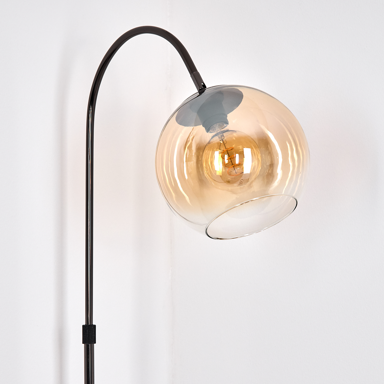 Eine moderne Stehlampe mit einem kugelförmigen, bernsteinfarbenen Glasschirm. Die Lampe hat ein schwarzes Metallgestell. Der Hintergrund ist eine weiße Wand.