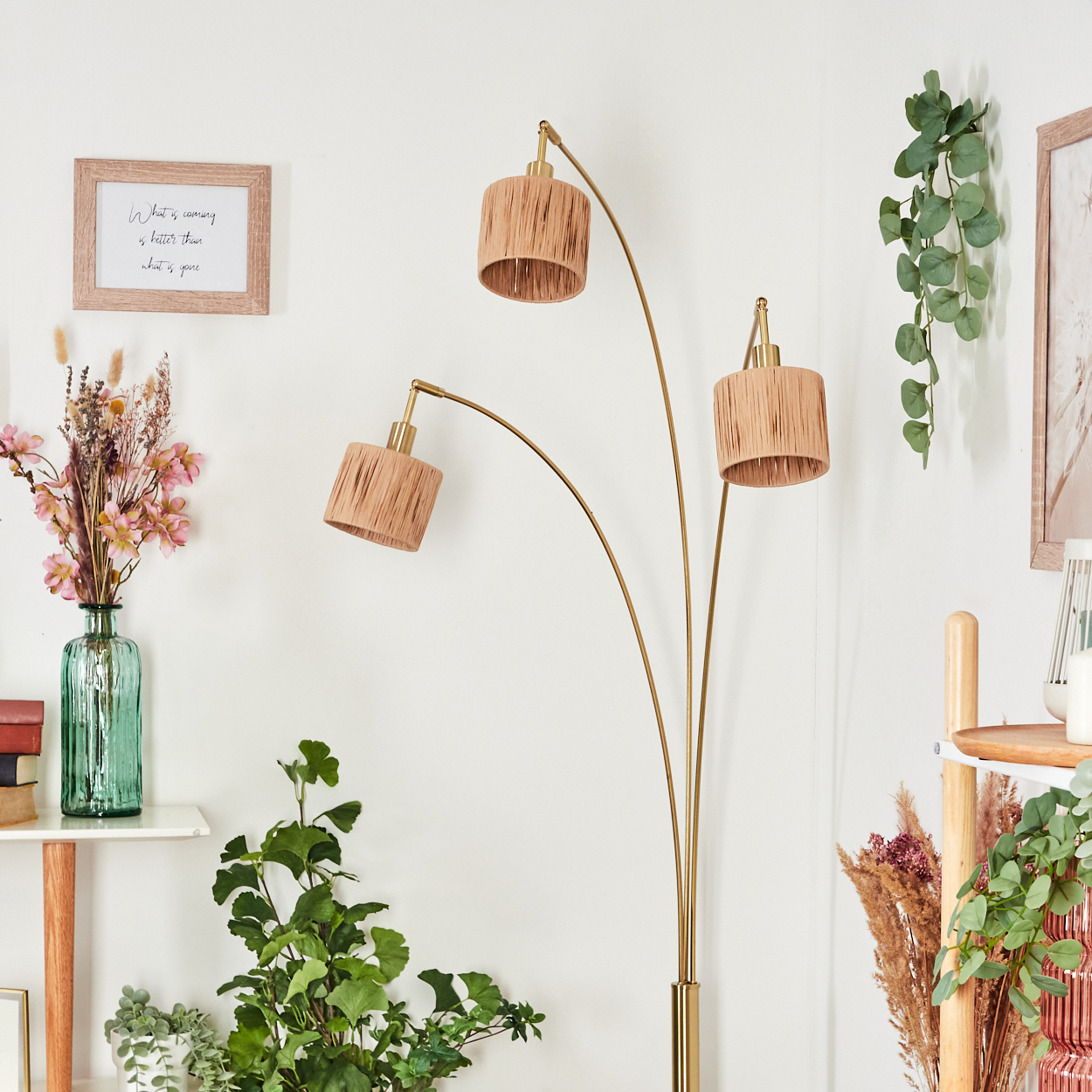 Eine Stehlampe mit drei beigefarbenen Schirmen, Pflanzen, gerahmter Kunst, einem Regal und einer Glasvase mit Blumen stehen vor einer weißen Wand.
