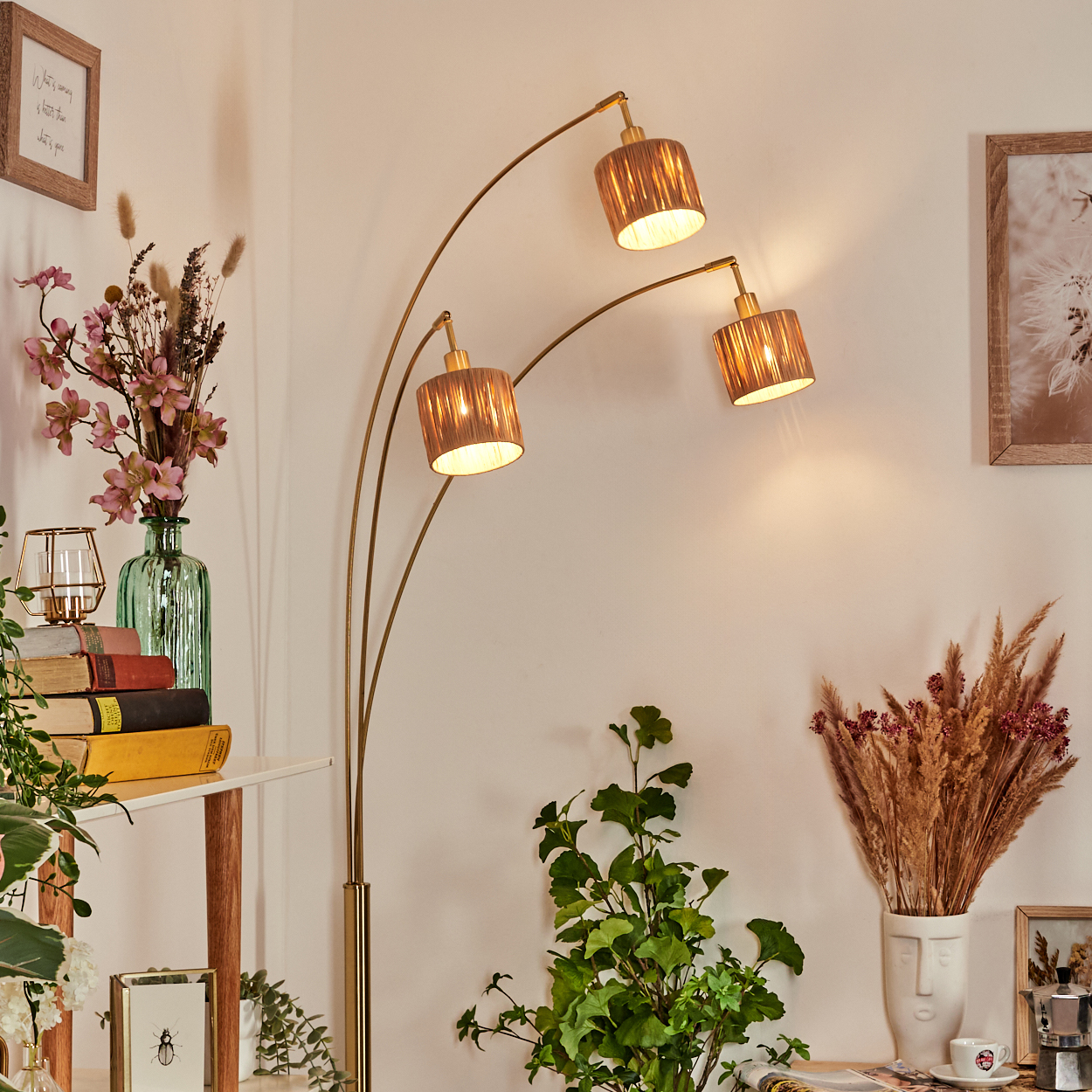 Eine Stehlampe mit drei Lampenschirmen beleuchtet einen Raum mit Pflanzen, Büchern und gerahmter Kunst. Beige Wände und ein weißes Regal schaffen eine gemütliche Atmosphäre.
