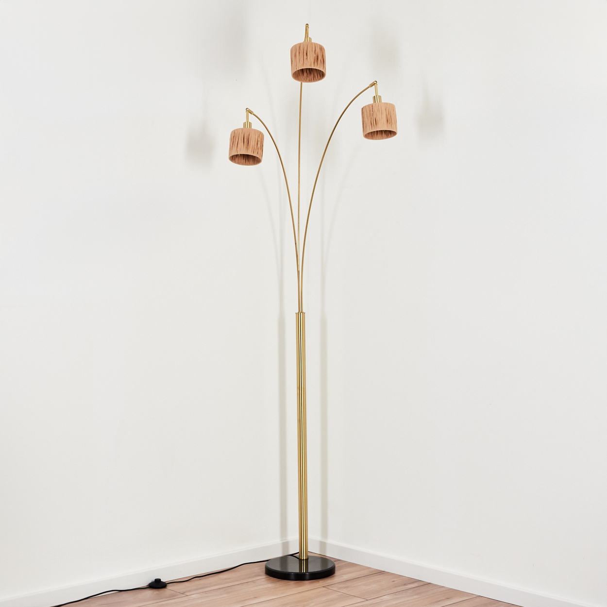Eine goldene Stehlampe mit drei Lampenschirmen. Die Lampenschirme sind beige. Der Sockel ist schwarz. Der Hintergrund ist eine weiße Wand und Holzfußboden.