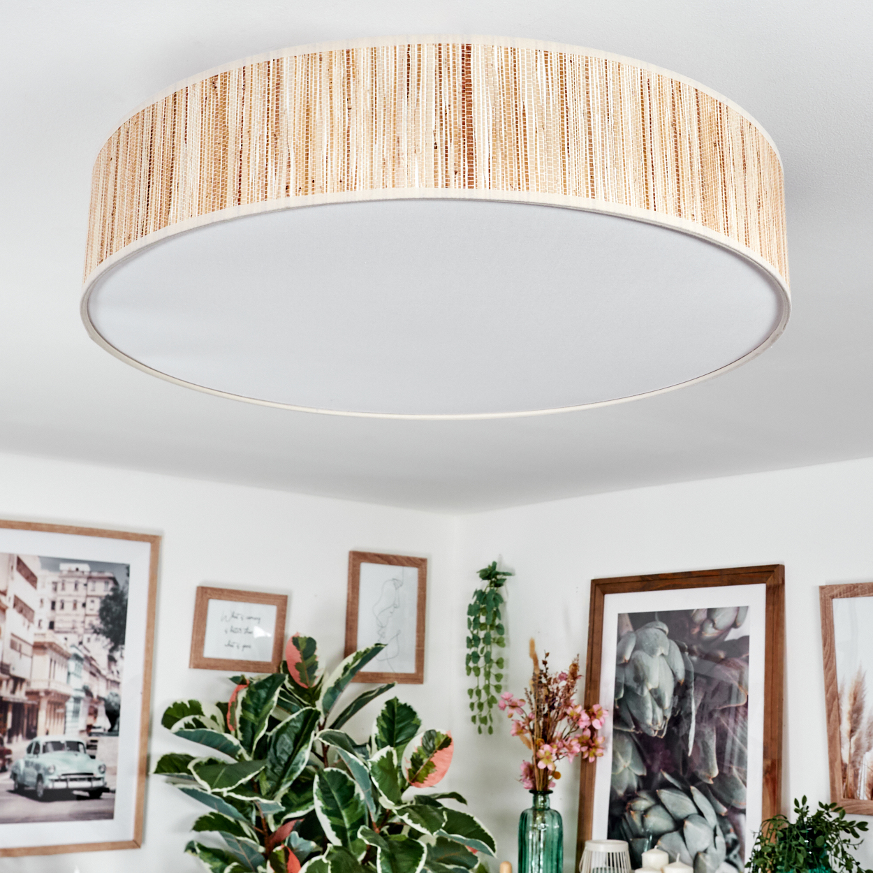 Eine runde Deckenlampe mit gewebtem Schirm. Darunter schmücken Bilder in Holzrahmen und Pflanzen die weiße Wand.