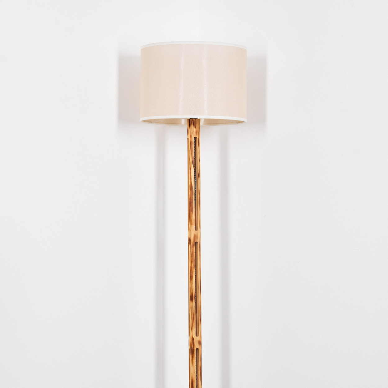Eine Stehlampe mit beigefarbenem Lampenschirm und einem bambusartigen Holzständer vor einer weißen Wand.