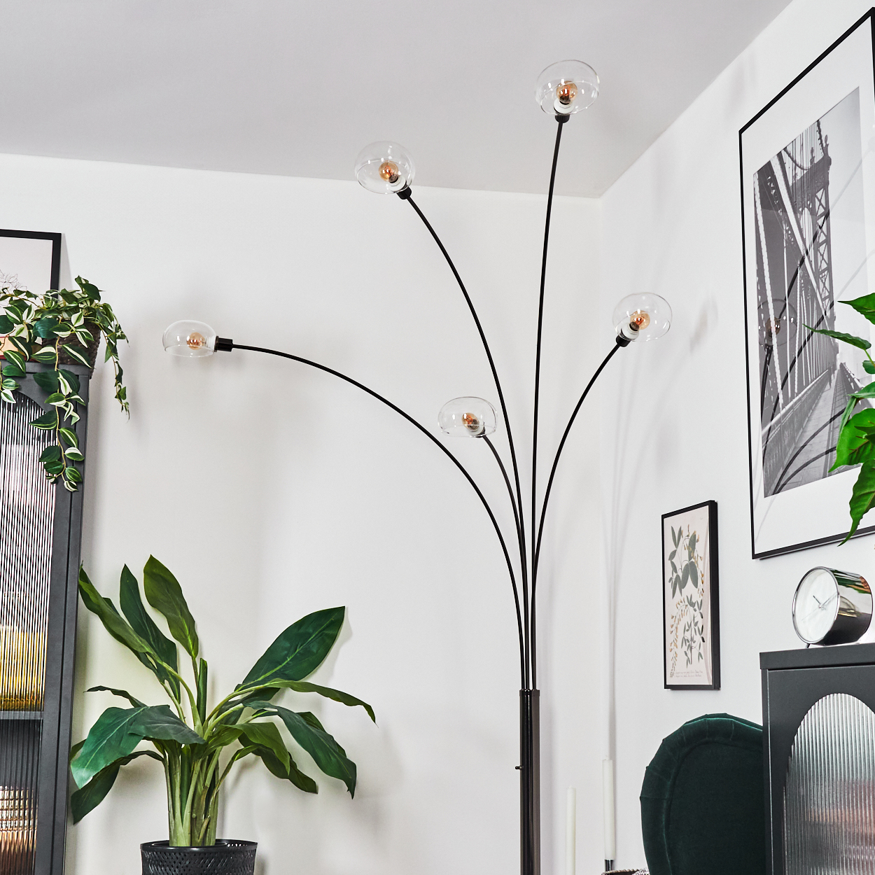 Eine moderne schwarze Stehlampe mit fünf Glasschirmen, neben einer großen Topfpflanze und gerahmten Kunstwerken in einem weißen Raum.