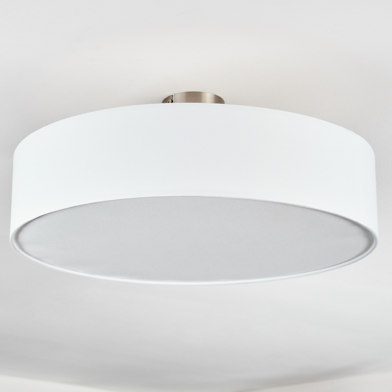 Eine weiße, kreisförmige Deckenlampe mit silbernem Sockel. Der Boden ist hellgrau. Der Hintergrund ist eine weiße Decke.