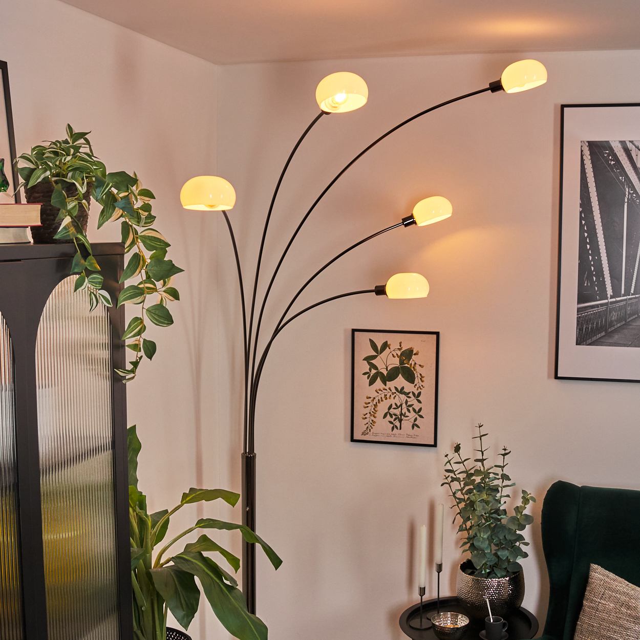 Ein modernes Wohnzimmer mit einer schwarzen Stehlampe mit fünf Glühbirnen. Pflanzen und Kunst schmücken den Raum. Im Hintergrund befindet sich ein grüner Samtstuhl.