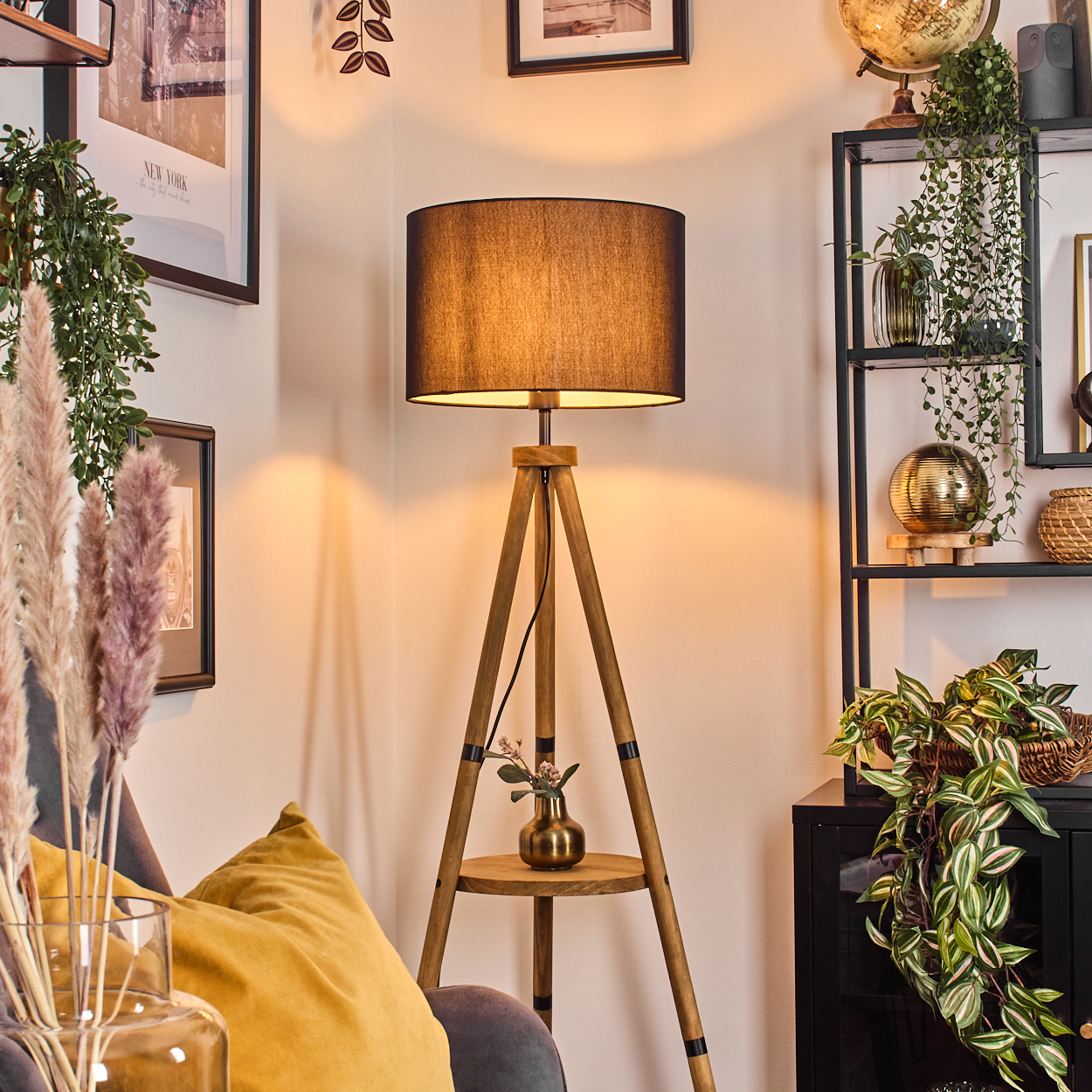 Eine Zimmerecke mit einer Holzstativlampe, einem gelben Kissen und einem schwarzen Bücherregal mit Pflanzen und Dekorationen.