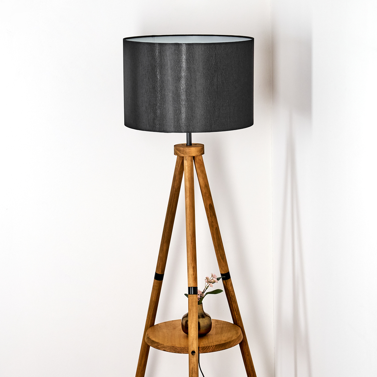 Eine hölzerne Stehlampe mit schwarzem Lampenschirm. Eine kleine Vase mit Blumen steht auf dem Regal. Der Hintergrund ist eine weiße Wand.
